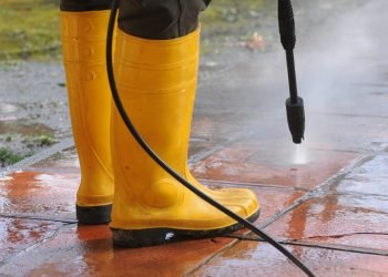 A person wearing yellow rubber boots with high-pressure water nozzle cleaning the dirt in the tiles